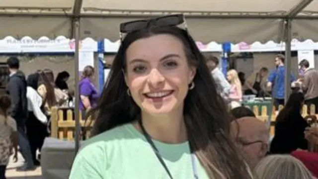 A woman with long brown hair standing under a marquee at the city food festival. She is wearing a green t-shirt and has sunglasses on the top of her head.