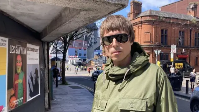 A man with short brown hair and sunglasses wearing a green jacket. He is stood on a pavement with buildings either side and the Principality Stadium visible behind him.