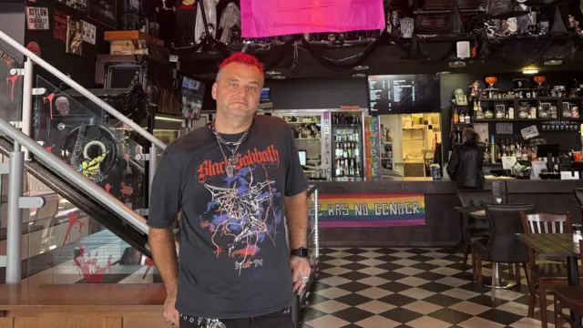 A man standing in front of a bar, he has red hair and is wearing a Black Sabbath top and black trousers.