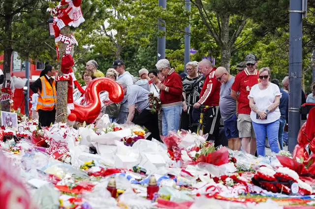 A large crowd of mourners stand looking at a grassy area completed obscured by flowers, scarves, balloons and football memorabilia
