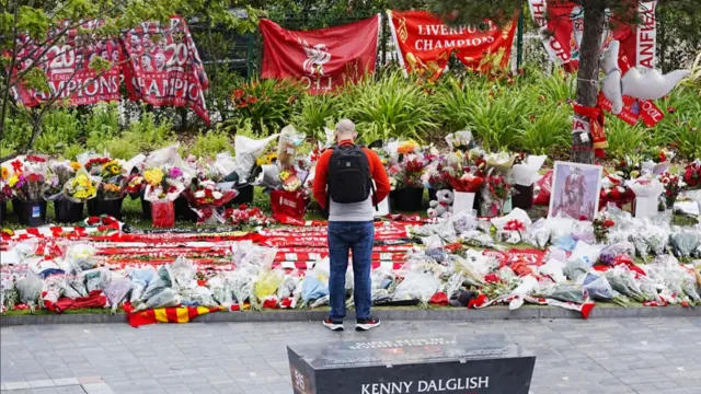 A man in long-sleeved red and white shirt stands in front of tributes outside Anfield stadium