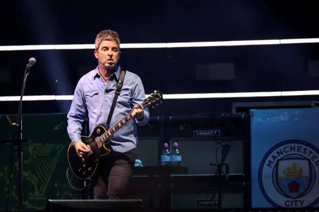 Noel Gallagher stands on stage holding an acoustic guitar, wearing a blue denim shirt and black jeans.
