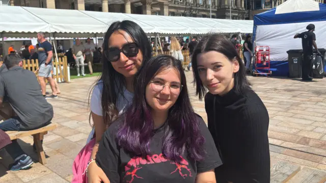 Three women who are in the middle of a city square. There are white marquees behind them. The women are young and all smiling.