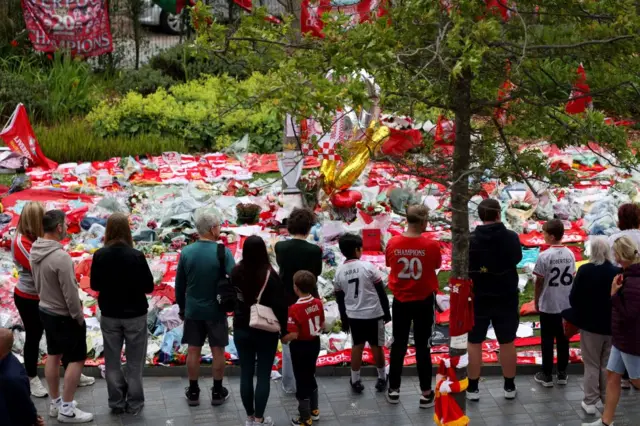 The back of fans looking over a tribute made to Diego Jots outside Anfield Stadium