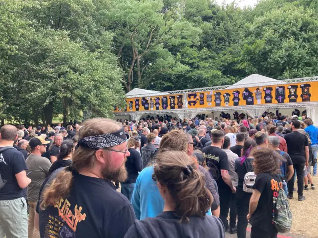 Hundreds of people are lined up in front of a long purple tent stand which houses merchandise. Along the top there are pieces of merch pinned up with prices next to them. The merch is largely black with purple writing