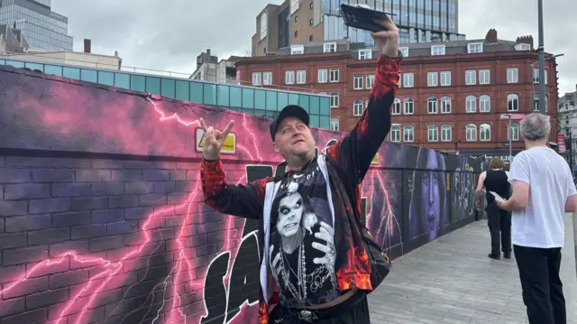 A man is taking a selfie and posing in front of a Black Sabbath mural on a long wall. He is wearing a black and white t-shirt with Ozzy Osbourne on it. He has a black jacket with red flames on it, a skull belt and a black cap