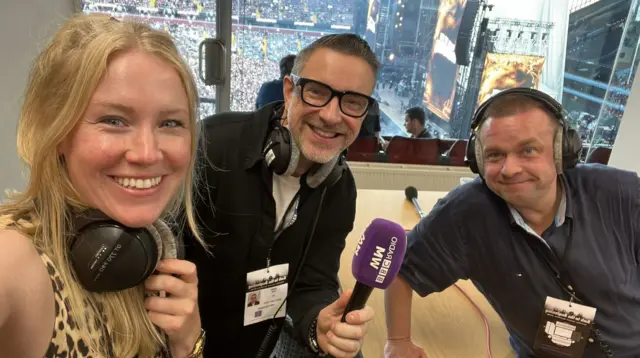 A woman next to two men, with the one in the middle holding a purple microphone