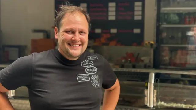 A man with brown hair is wearing a black t-shirt and has his hands on his hips. He is standing in front of a fast food van