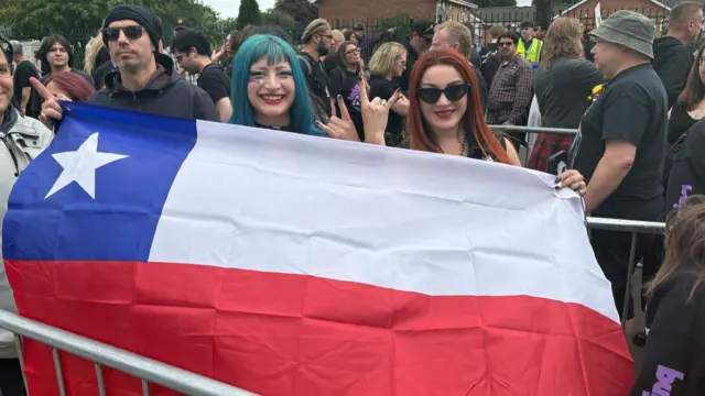A woman with blue hair stands next to a woman with red hair. They are holding the Chile flag. their ands are up and they are doing the rock hand symbol