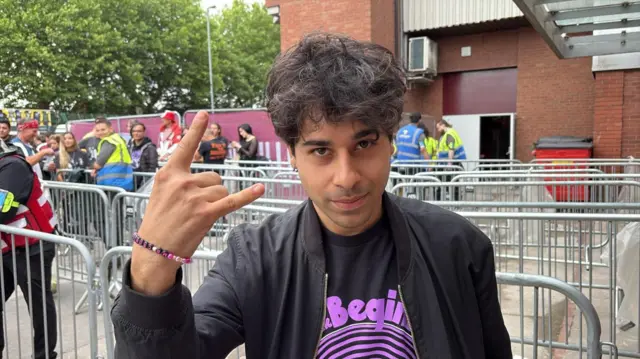 A man with curly dark brown hair is wearing a black jacket and a black t-shirt with purple writing and a purple swirl on it. He has one hand in he air and is doing the rock hand symbol