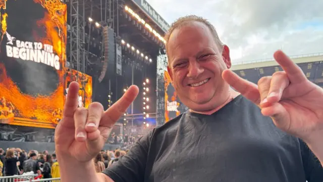 A man makes the horns of rock symbol with both hands in front of a stage with lights on and around it.He has short dark brown hair and smiles into the camera