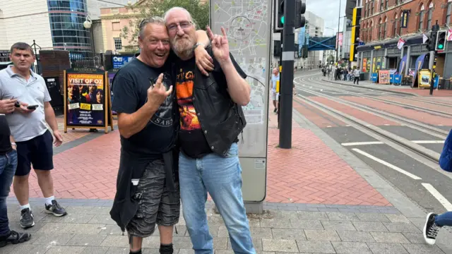 Two men have their arms around each other and have their hands in the air with the rock hand symbol. They are both wearing black band t-shirts