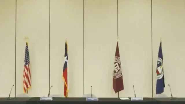 A table covered in black cloth holding five mics, four flags and flagpoles in the background in front of a white wall