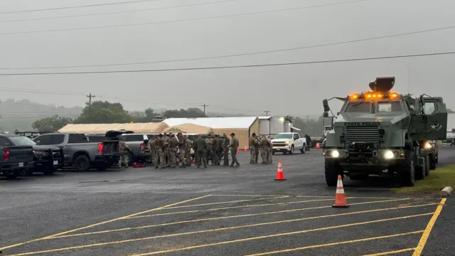 Wide shot of large parking lot with military vehicle parked to the right of the frame as a large group of men in military apparel stands behind parked cars to the left
