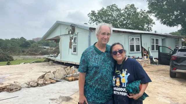 An elderly man (L) in a short sleeved, patterned polo with his left arm behind a shorter woman (L) in a Bluey dark blue shirt, a damaged blue and green house behind them