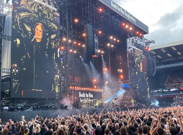 Ozzy Osbourne on the big screen with people in the crowd with their arms in the air.
