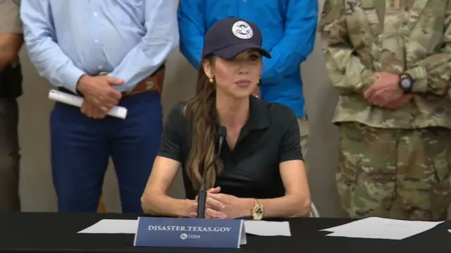 Kristi Noem sits at table covered in black cloth wearing a short-sleeved polo shirt and a cap with the DHS insigna, several officials standing behind her