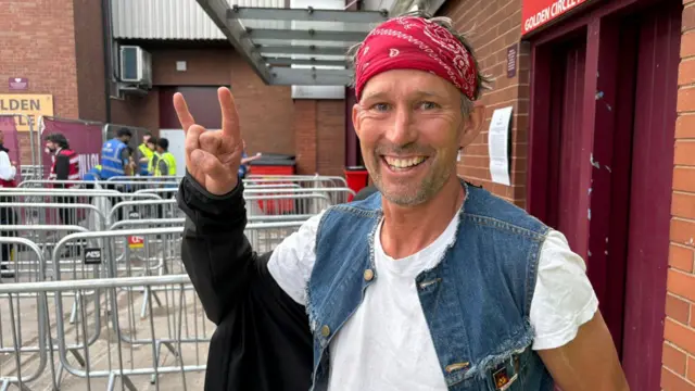 A man with grey hair is wearing a red bandanna, white t-shirt and denim gilet jacket. He is queueing outside a stadium. He has one arms in the air doing the rock hand sign