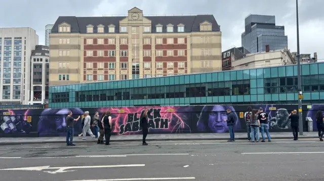 A long wall in Birmingham with a Black Sabbath mural on it. There are people standing in front of it taking pictures and selfies