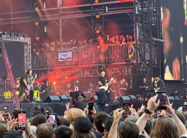 A man on stage with a guitar and some people in the crowd holding up their mobiles.