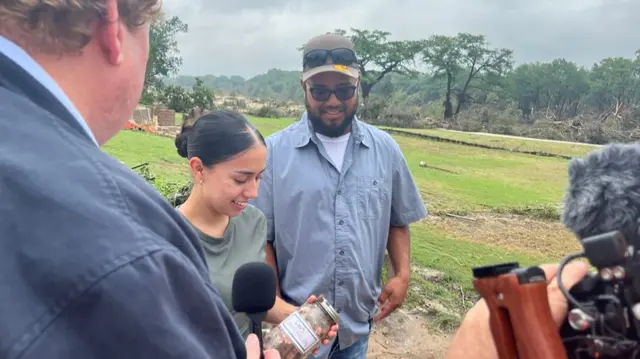 BBC reporter (L) with his back to the camera hold a mic up to a young woman smiling down at a jar filled with coins with the label 'Leo's survival kit'. A man (C) in short-sleeved shirt and cap also looks down at the jar