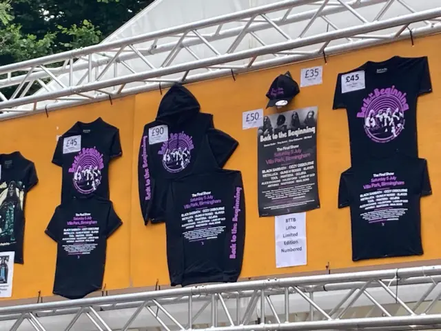 Black and purple merchandise hung up on a stand. there are prices next to each piece, which include t-shirts, hoodies and caps.