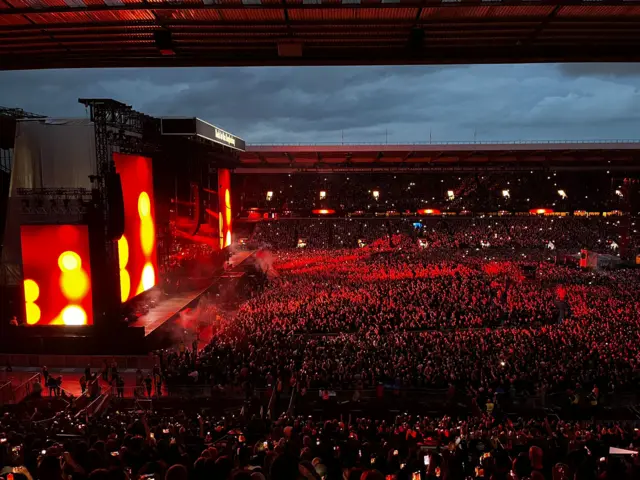 A stage lights up in front of a concert crowd of thousands as red light bathes the crowd