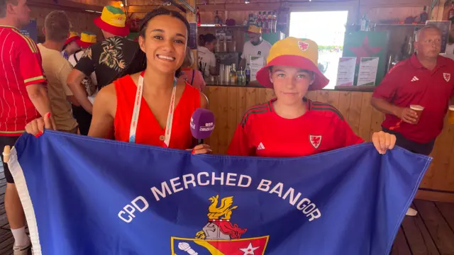 Newsround presenter Nina with young fan Ellie, wearing a bucket hat and holding up a flag for a local Bangor team