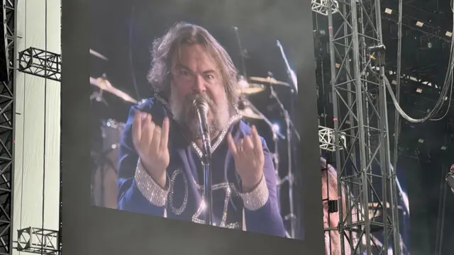 Jack Black has long grey and brown hair and is on screen in a stadium. He is singing into a microphone and has his hands in the "rock" symbol. He is wearing a blue top decorated with rhinestones that reads "ozzy"