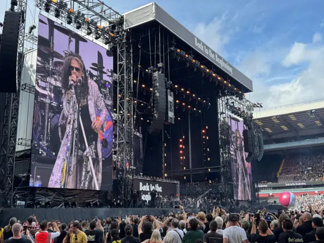 A wide shot showing the audience on the ground and the stage, with big screens to the left and right of it.