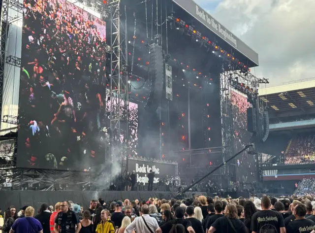 Big screens either side of the stage showing the crowd. Those in the crowd are also visible on the ground.