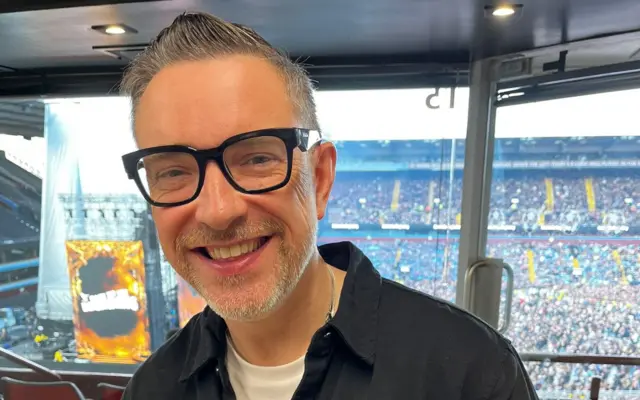 A man with glasses is smiling at the camera. Stands at Villa Park are behind him.