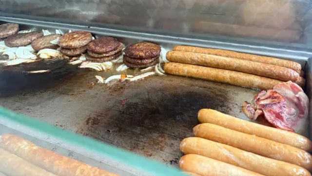 Burgers, sausages, bacon and onions are frying on a large industrial cooker inside a food van