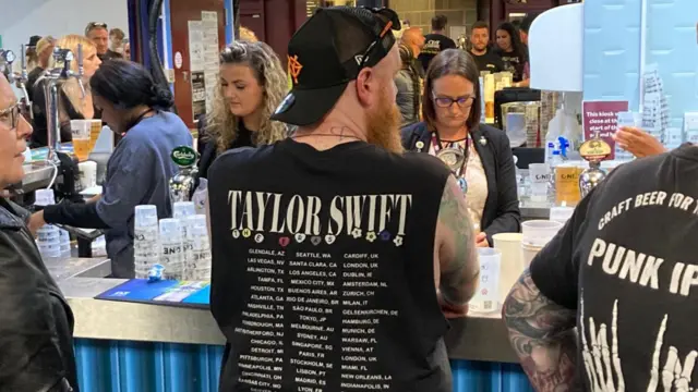 The back of a man standing at a bar during a concert. He has a black t-shirt on that reads "taylor swift, the eras tour"