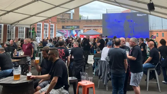 Dozens of people are sitting under an outdoor gazebo, drinking pints and talking. There's a large screen mounted behind them
