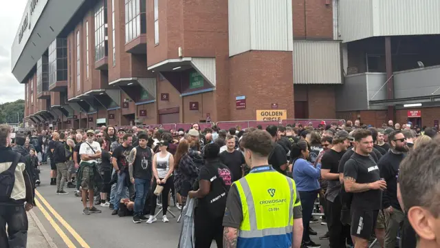 Thousands of people are standing outside a large football stadium. There is someone in a fluorescent jacket watching them