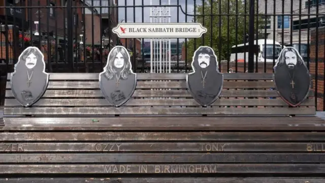 The bench is brown and has four black and white photos of the Black Sabbath members on it. there is a sign behind it on railings that says Black Sabbath bridge