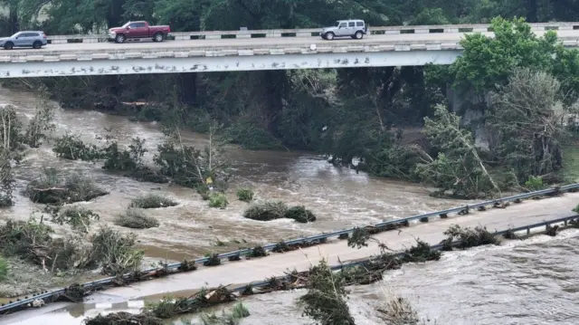 A drone view shows debris on a road, as a result of flash flooding, in Comfort, Texas as three pick up trucks drive down a bridge road to the top-left of the one submerged in flood waters