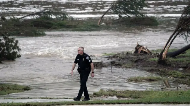 A Kerrville PD officer in uniform walks on the pavement, the road behind him submerged by flood water, some patches of grass and bent trees behind him
