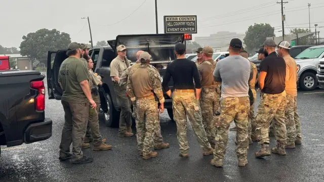 Members of the Kerrville Police Department wearing camo trousers and shirts wait around the back of a grey pick up truck in a parking lot