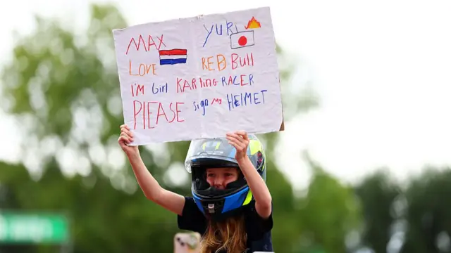 A young go-karter fan holds a sign asking Max Verstappen and Yuki Tsunoda to sign her helmet