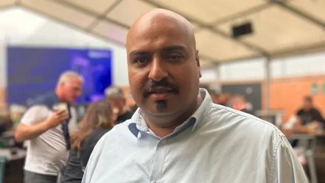 Kiran is bald and is wearing a light blue shirt. Behind him are punters inside the pub and a large screen mounted behind him
