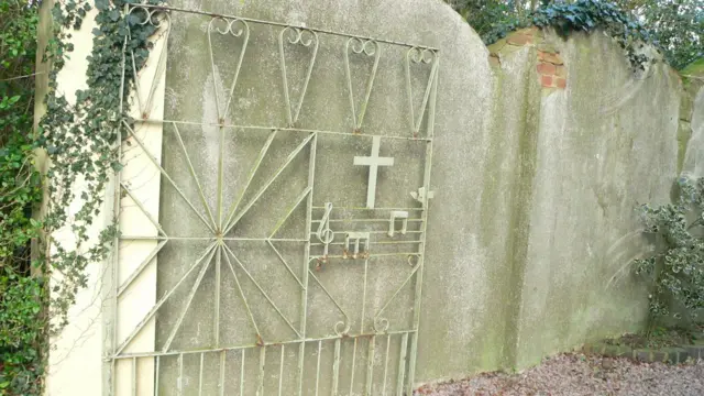 A white gate now faded slightly green with age. Part of the metal is shaped into a cross, with music notes
