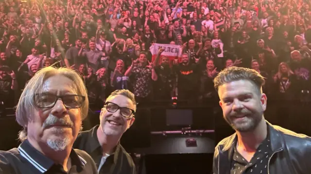 Three men are standing on a stage with their backs to the crowd, taking a selfie. There are hundreds of fans on seats in the background with their arms in the air, making rock symbols with their hands