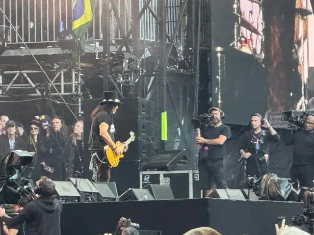 A man with a hat and a guitar is performing on stage. A camera operator is on the right of the photo.