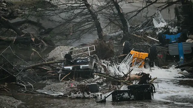 A man in a black top walks through flood water as he passes overturned cars, tractors and trees, water gushing along the road to the right of the frame