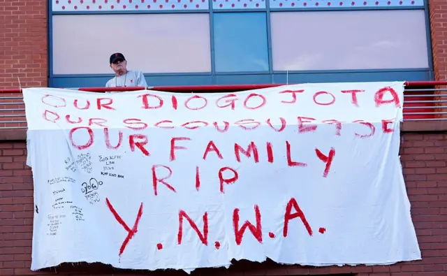 A man holds a white sheet at Anfield which says 'Our Diogo Jota. Our Scousuesse, Our Family, RIP, YNWA'