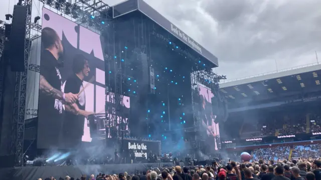 People are standing below a stage watching a band perform. Writing on the stage reads "Back to the Beginning"