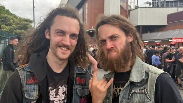 Two men with long hair are wearing black band t-shirts and denim gilet style jackets. One has long facial hair and is holding up his hand, doing the rock hand symbol