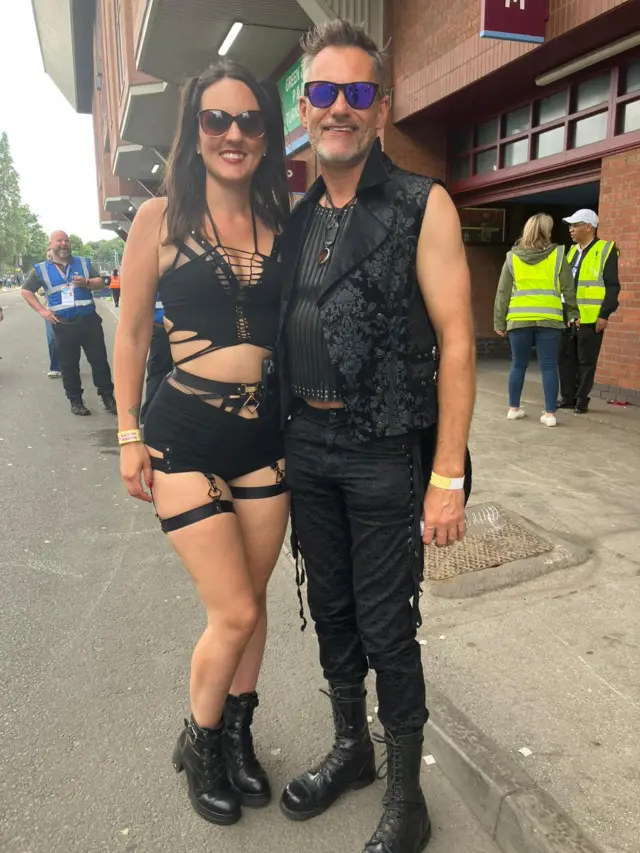 A man and woman standing outside a stadium. The woman has a black crop top and shorts on with ripped pieces of material, and black boots. the man is in all black with high black boots and a decorative gilet